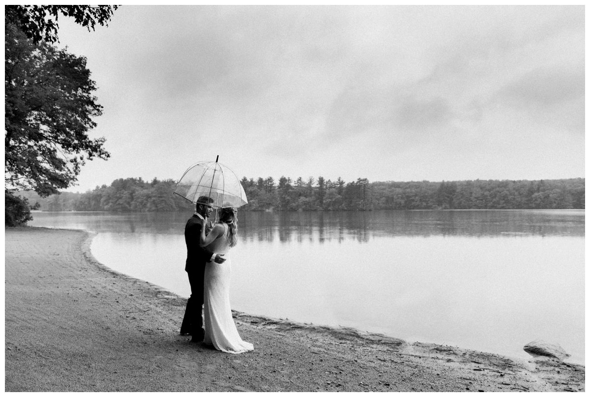 black and white photo of bride and groom embracing under umbrella on the shore