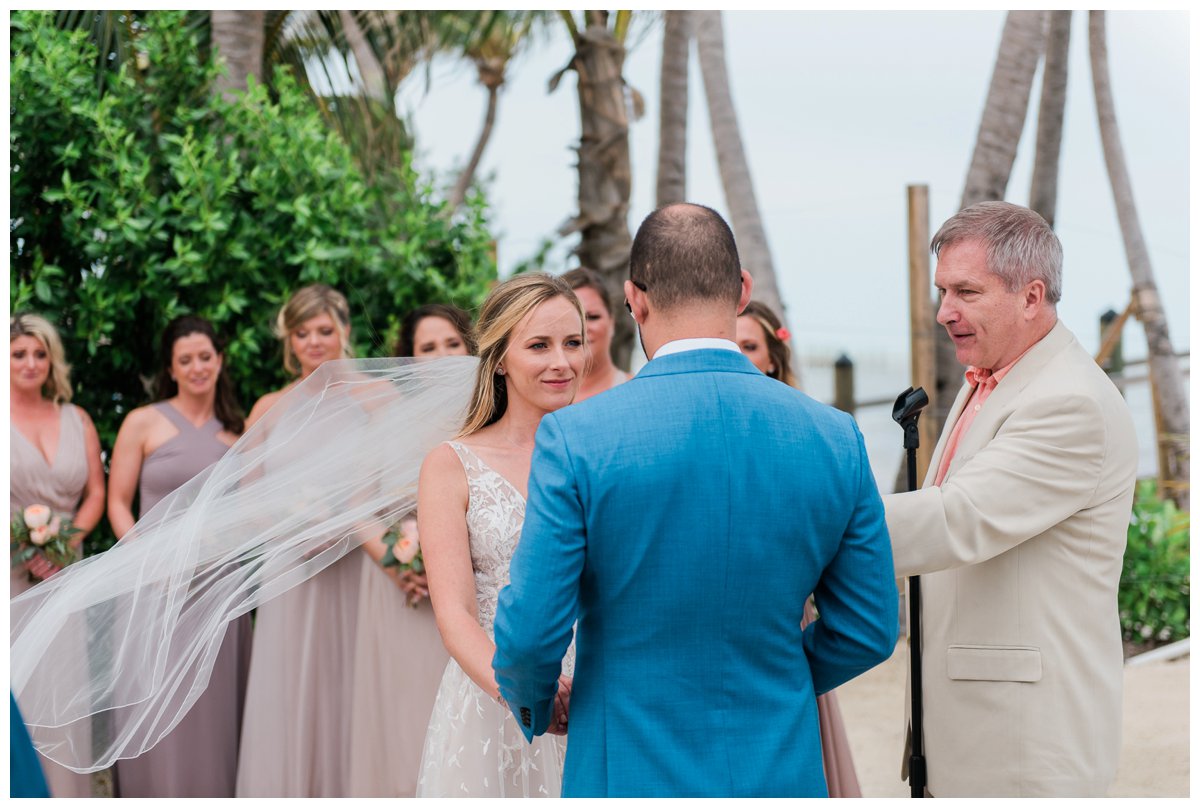 the bride's veil blows in the wind on the beach for her tropical wedding
